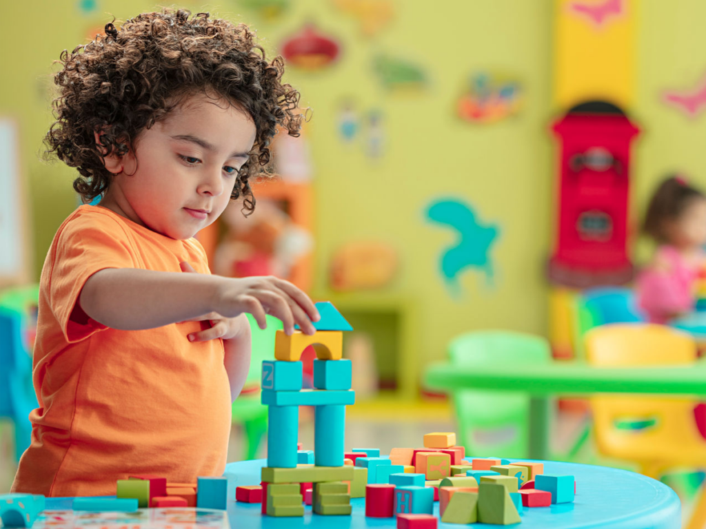 A young child wearing an orange shirt is deeply focused while stacking colorful wooden blocks to build a structure. The background shows a vibrant playroom with cheerful decorations, including wall art and brightly colored furniture. This scene highlights the joy of imaginative play, which can be enhanced with pre-loved toys that are both sustainable and fun.