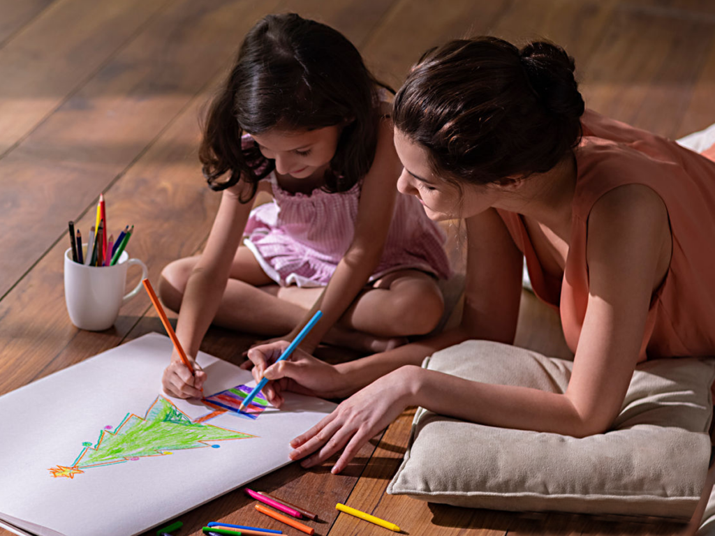 A mother and daughter sitting on the floor, drawing a colorful Christmas tree together on a large sheet of paper, surrounded by colored pencils and a mug filled with them.